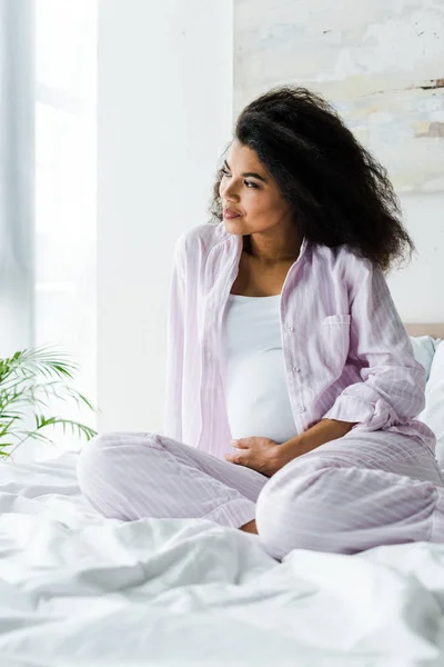 Sonriente joven embarazada africana americana mujer sentada en la cama - foto de stock
