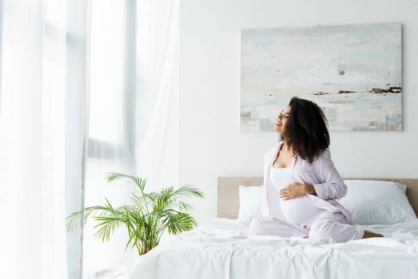 Happy young pregnant african american woman sitting on bed and touching belly — Stock Photo