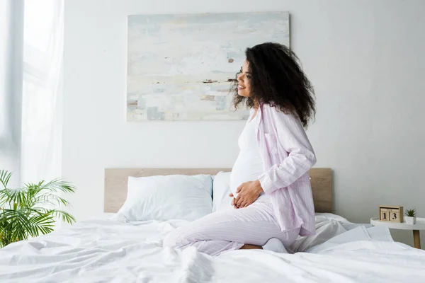 Jovem e alegre mulher americana africana grávida sentada na cama e tocando barriga — Fotografia de Stock