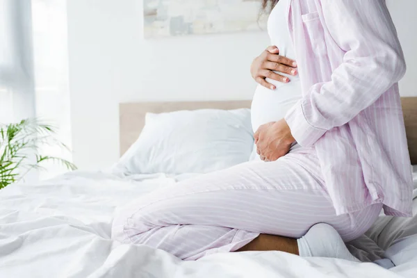 Vista recortada de mujer afroamericana embarazada sentada en la cama y tocando el vientre - foto de stock