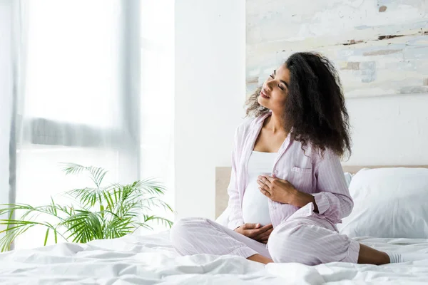 Gai jeune femme enceinte afro-américaine avec les yeux fermés assis sur le lit et toucher le ventre — Photo de stock