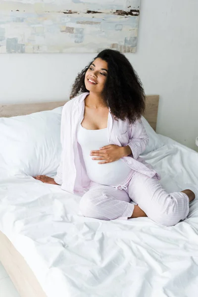 Positivo joven embarazada africana americana mujer sentada en la cama y tocando vientre - foto de stock