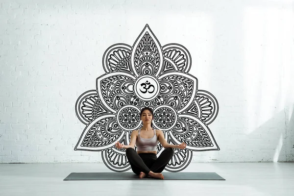 Calm asian woman with closed eyes sitting on yoga mat near mandala ornament — Stock Photo