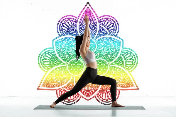 Asian woman practicing yoga on yoga mat near colorful mandala ornament on white — Stock Photo