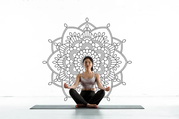 Спокойная тайская женщина практикующая йогу на коврике для йоги рядом с мандаловым орнаментом на белом — стоковое фото
