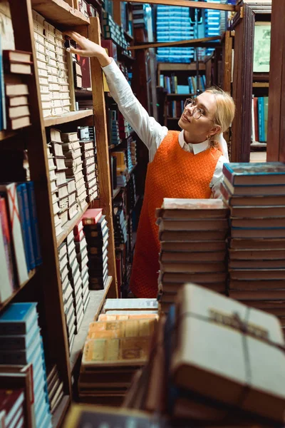Bella e bionda donna in abito arancione e bicchieri che tengono libro in biblioteca — Foto stock