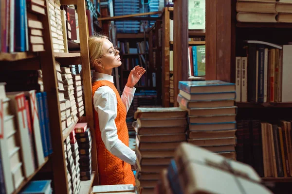 Mujer bonita y rubia en vestido naranja y gafas de pie en la biblioteca - foto de stock