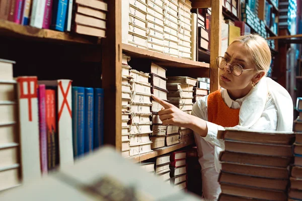 Mulher bonita e loira em óculos apontando com o dedo para o livro na biblioteca — Fotografia de Stock