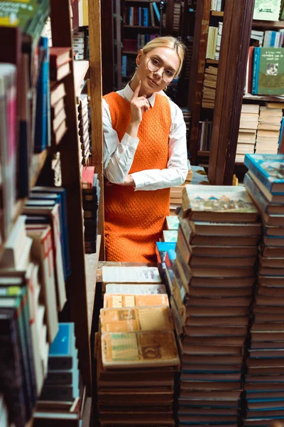 Mulher bonita e loira em óculos e vestido laranja olhando para longe na biblioteca — Fotografia de Stock
