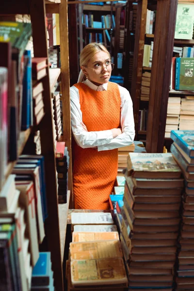 Mulher bonita e triste em óculos e vestido laranja com braços cruzados olhando para longe na biblioteca — Fotografia de Stock