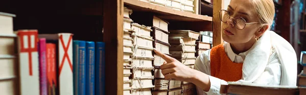 Панорамный снимок красивой блондинки в очках, указывающей пальцем на книгу в библиотеке — стоковое фото
