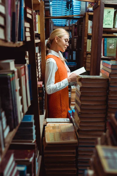 Вид сбоку симпатичной блондинки в очках и оранжевом платье читающей книги в библиотеке — стоковое фото
