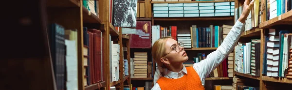 Panoramic shot of pretty and blonde woman in glasses holding book in library — Stock Photo