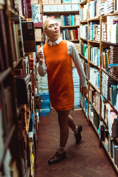 Belle et blonde femme en lunettes et robe orange regardant loin dans la bibliothèque — Photo de stock