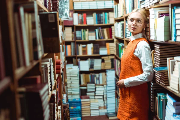 Schöne, blonde Frau mit Brille und orangefarbenem Kleid, die in der Bibliothek steht und wegschaut — Stockfoto