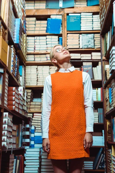 Blonde et belle femme en lunettes et robe orange regardant vers le haut dans la bibliothèque — Photo de stock
