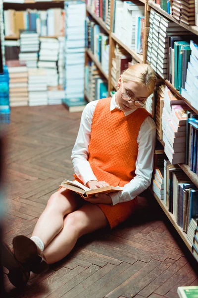 Bionda e bella donna in occhiali e abito arancione seduta sul pavimento e libro di lettura in biblioteca — Foto stock