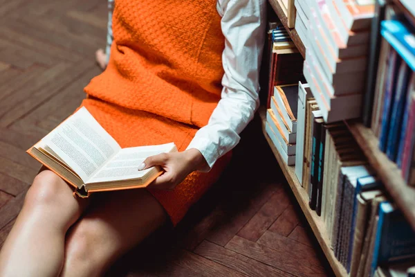Vue recadrée de la femme en robe orange assise sur le sol en bois et tenant livre — Photo de stock