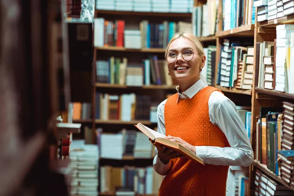 Sorrindo e bela mulher em vestido laranja segurando livro e olhando para longe na biblioteca — Fotografia de Stock
