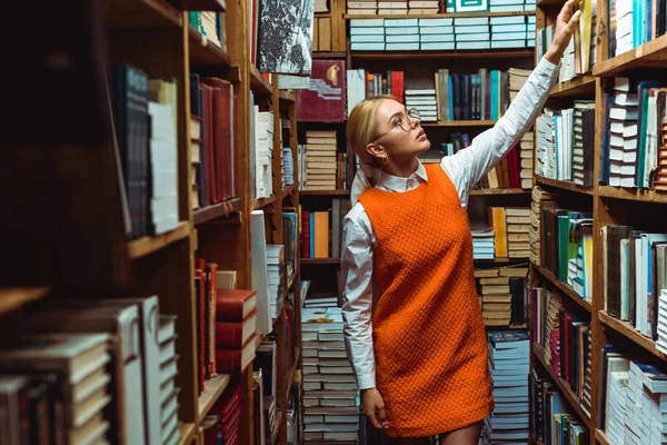 Mulher loira e bonita em vestido laranja segurando livro e olhando para longe na biblioteca — Fotografia de Stock