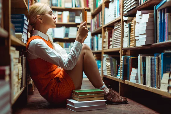 Hermosa y rubia mujer en gafas lectura libro y sentado en el suelo en la biblioteca - foto de stock