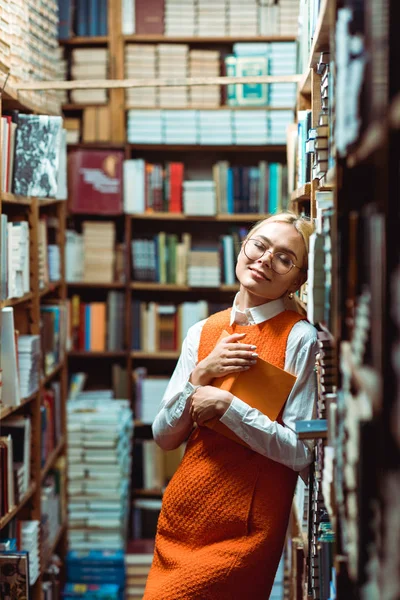 Belle femme blonde avec les yeux fermés dans des lunettes tenant livre dans la bibliothèque — Photo de stock