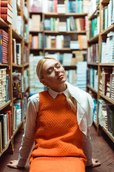 Beautiful and blonde woman with closed eyes in glasses sitting on floor in library — Stock Photo