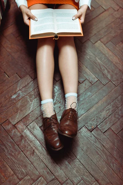 Vista cortada da mulher sentada no chão de madeira e livro segurando — Fotografia de Stock