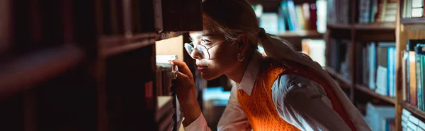 Plan panoramique de jolie femme en lunettes regardant loin dans la bibliothèque — Photo de stock