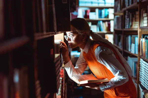 Bonita mujer en gafas mirando hacia otro lado y sosteniendo el libro en la biblioteca - foto de stock