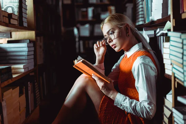 Bella e bionda donna in occhiali seduta e lettura libro in biblioteca — Foto stock