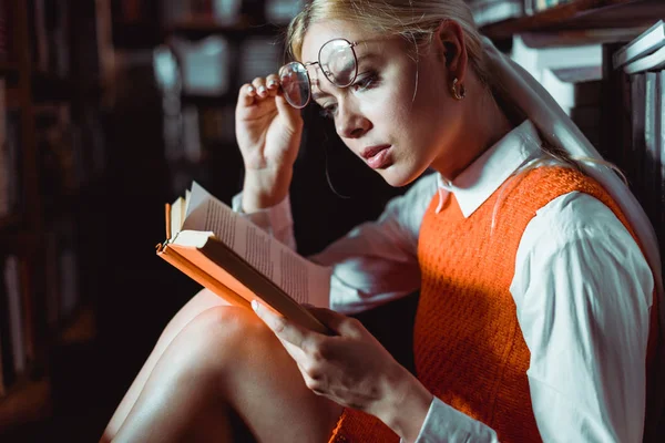 Beautiful and blonde woman holding glasses and reading book in library — Stock Photo