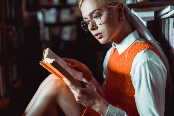 Schöne und blonde Frau mit Brille liest Buch in Bibliothek — Stockfoto