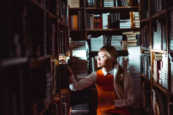 Belle et blonde femme en lunettes et robe orange regardant loin dans la bibliothèque — Photo de stock