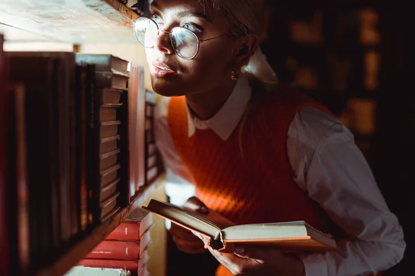 Mulher bonita em óculos segurando livro e olhando para longe na biblioteca — Fotografia de Stock