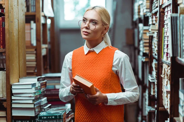 Hermosa y rubia mujer en gafas sosteniendo libro y mirando hacia otro lado en la biblioteca - foto de stock