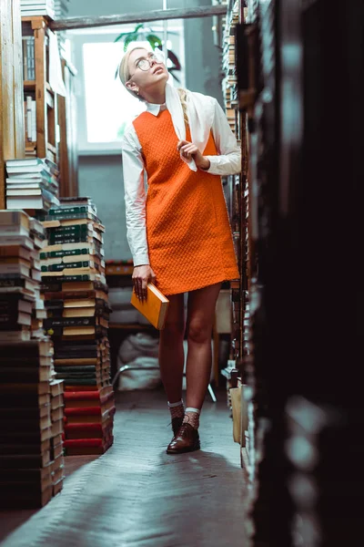 Mulher bonita e loira em óculos e vestido laranja segurando livro e olhando para cima na biblioteca — Fotografia de Stock