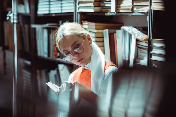 Schöne und blonde Frau mit Brille liest Buch in Bibliothek — Stockfoto