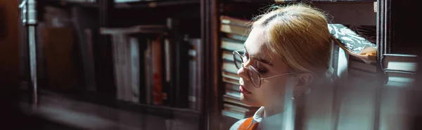 Panoramic shot of blonde and attractive woman in glasses in library — Stock Photo