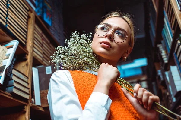 Low angle view of woman in glasses holding white flowers and looking away in library — Stock Photo