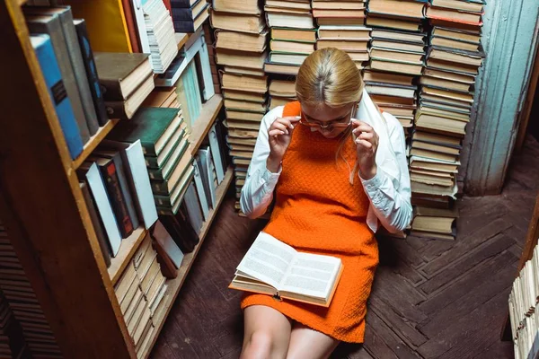 Hermosa mujer en vestido naranja sentado en el suelo y libro de lectura en la biblioteca - foto de stock