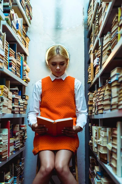 Mulher bonita e loira em vestido laranja leitura livro na biblioteca — Fotografia de Stock