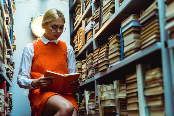 Schöne und blonde Frau in orangefarbenem Kleid und Brille liest Buch in der Bibliothek — Stockfoto