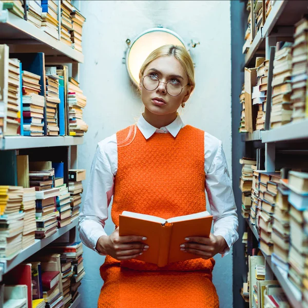 Mulher bonita e pensativa em vestido laranja segurando livro e olhando para cima na biblioteca — Fotografia de Stock
