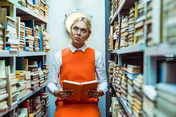 Mujer hermosa y pensativa en vestido naranja sosteniendo libro y mirando hacia arriba en la biblioteca - foto de stock