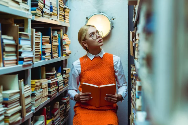 Beautiful and blonde woman in orange dress holding book and looking up in library — Stock Photo