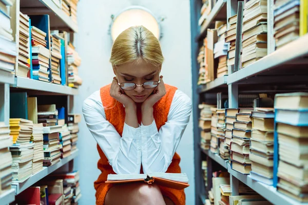 Beautiful and blonde woman in orange dress and glasses reading book in library — Stock Photo