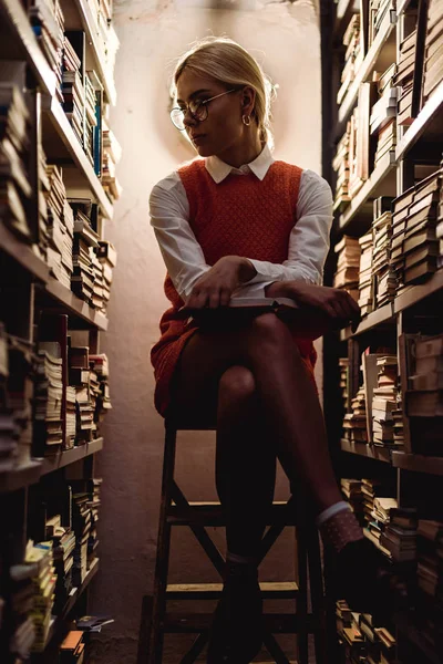 Mulher bonita e loira em vestido laranja sentado na escada e segurando livro na biblioteca — Fotografia de Stock