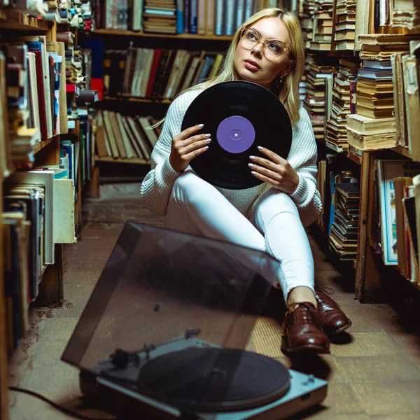 Beautiful and blonde woman sitting on floor and holding vinyl in library — Stock Photo