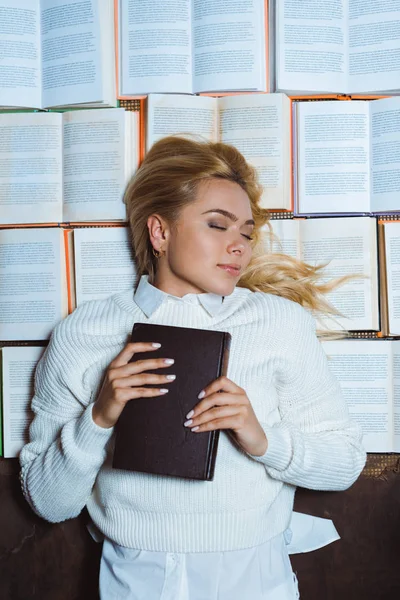 Atractiva y rubia mujer con los ojos cerrados mintiendo y sosteniendo libro - foto de stock